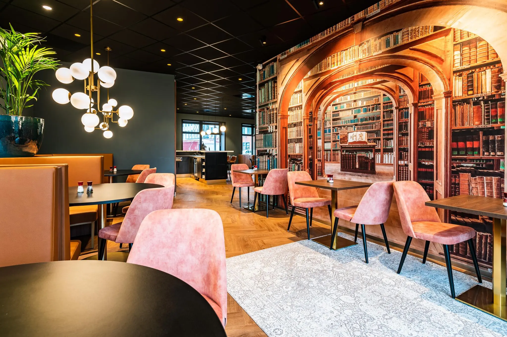 Modern restaurantinterieur aan de George Hintzenweg met roze stoelen, leren zitbanken en een opvallende wanddecoratie met een bibliotheekprint.